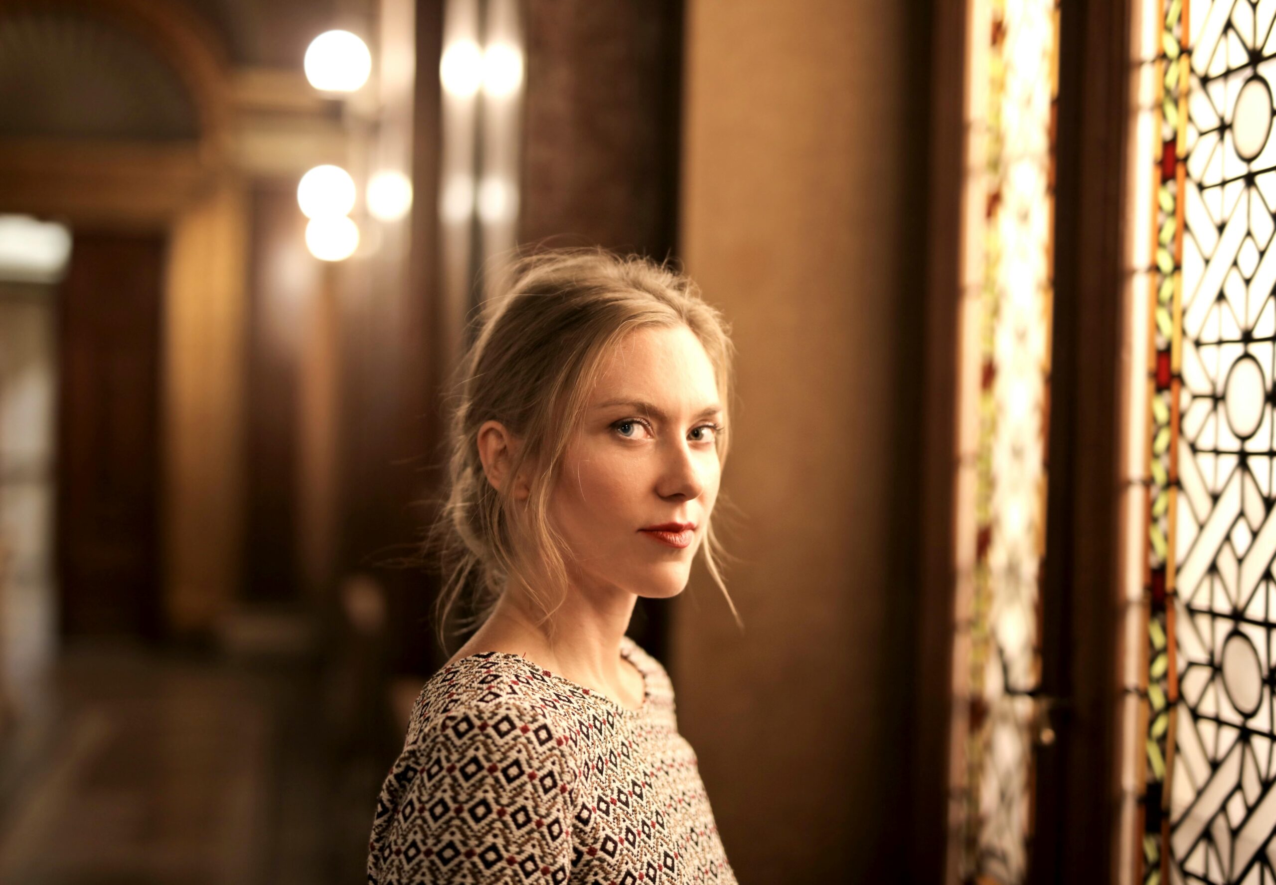 Portrait of a woman in an elegant, warm-toned room with stained glass window. Artistic and serene.